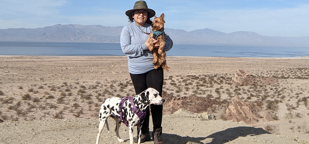 A womam wearing a blue jacket stands in the center of the desert holding a small brown dog. A dalmation is to her left.
