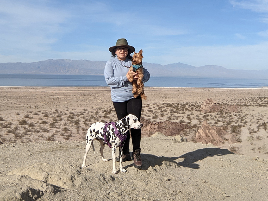 A womam wearing a blue jacket stands in the center of the desert holding a small brown dog. A dalmation is to her left.